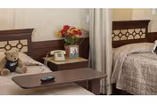 A nursing home bedroom with two beds featuring matching beige bedspreads. Between the beds is a nightstand with a telephone, a framed photo, and a vase of flowers. A teddy bear is sitting on one of the beds, and a remote control is on a brown overbed table in the foreground.