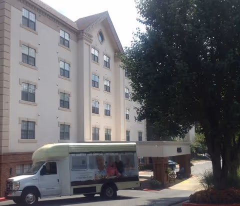 Exterior front of a multi-story retirement community building with a shuttle van parked under a covered entrance and trees along the driveway.