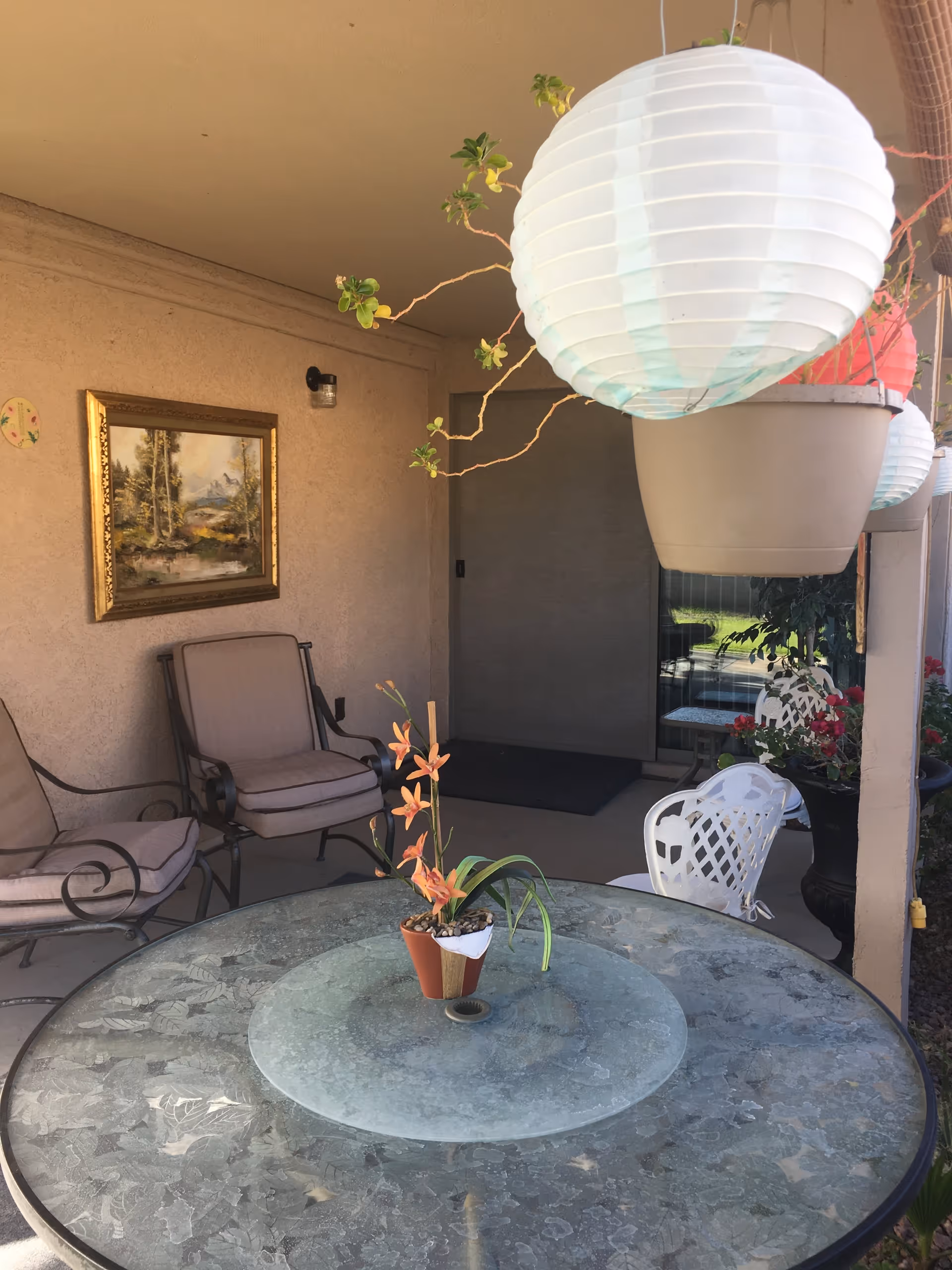 Covered outdoor patio with a round glass table and potted plant centerpiece, cushioned chairs, hanging paper lanterns, and a framed painting on the wall.