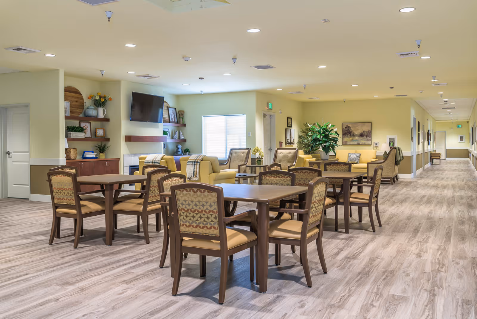 A spacious and well-lit common area in a senior living facility with several wooden tables and cushioned chairs arranged for group seating. The room features a seating area with yellow sofas and armchairs, a wall-mounted flat-screen TV, decorative shelves with plants and artwork, and a long hallway extending to the right. The flooring is light wood, and the walls are painted a soft yellow with white trim.