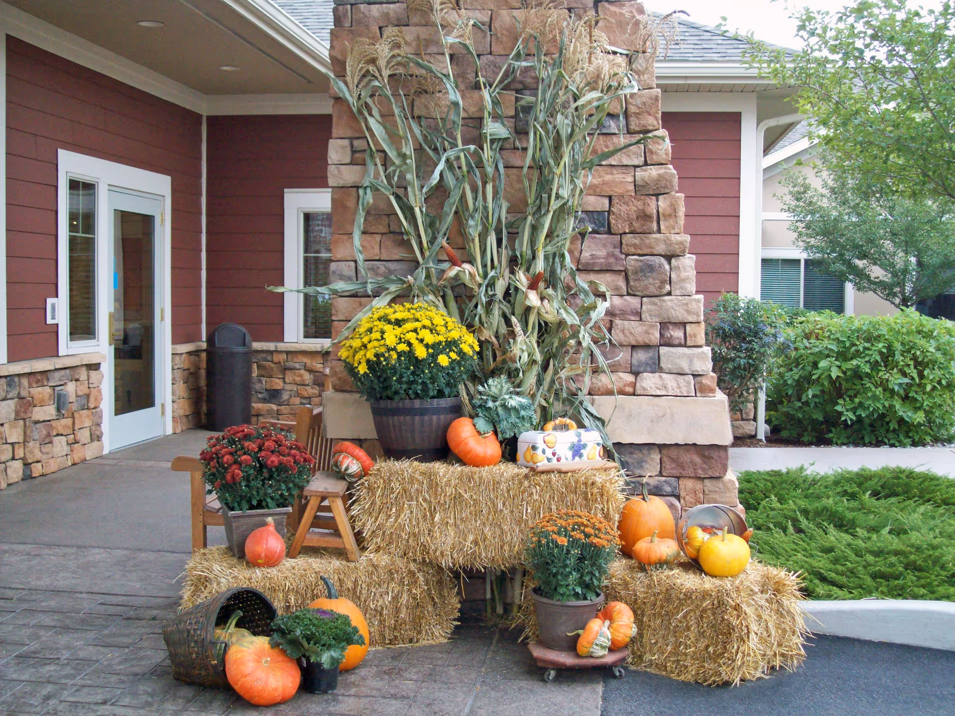 Outdoor autumn-themed decoration with hay bales, pumpkins, potted chrysanthemums, and dried corn stalks arranged in front of a stone pillar near the entrance of a building with red siding and white trim.