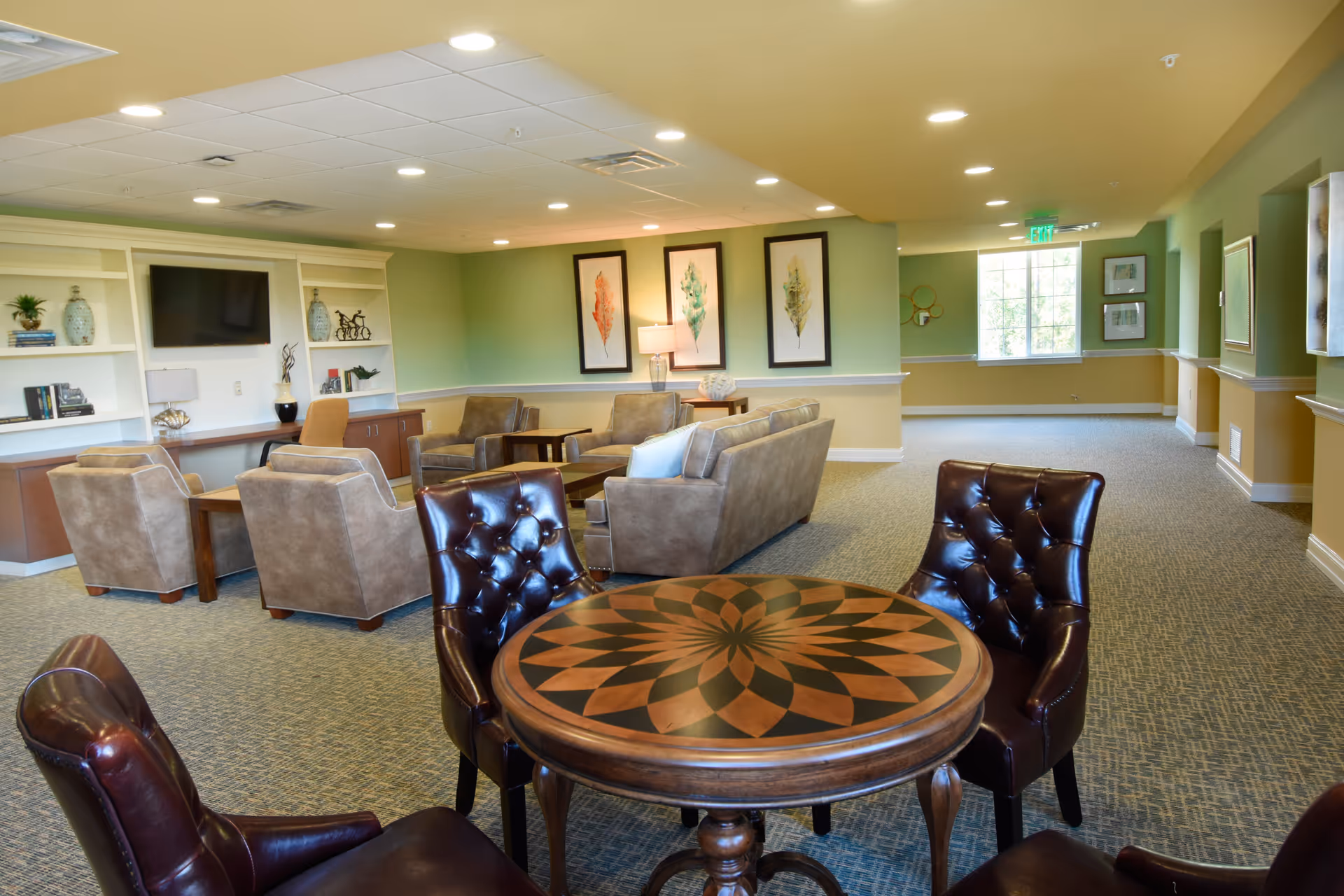 A spacious communal lounge with upholstered sofas and chairs, a decorative round table with leather chairs, a wall-mounted TV and framed artwork.