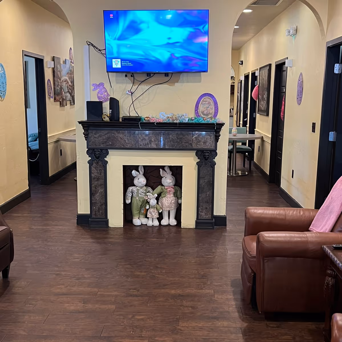 Interior view of a senior living facility hallway with a mounted flat-screen TV above a decorative fireplace mantel. The fireplace opening contains three stuffed bunny dolls dressed in clothes. The walls are decorated with Easter-themed decorations and artwork. There are doorways on both sides of the hallway, a small table with chairs in the background, and a brown leather armchair with a pink blanket on the right side.