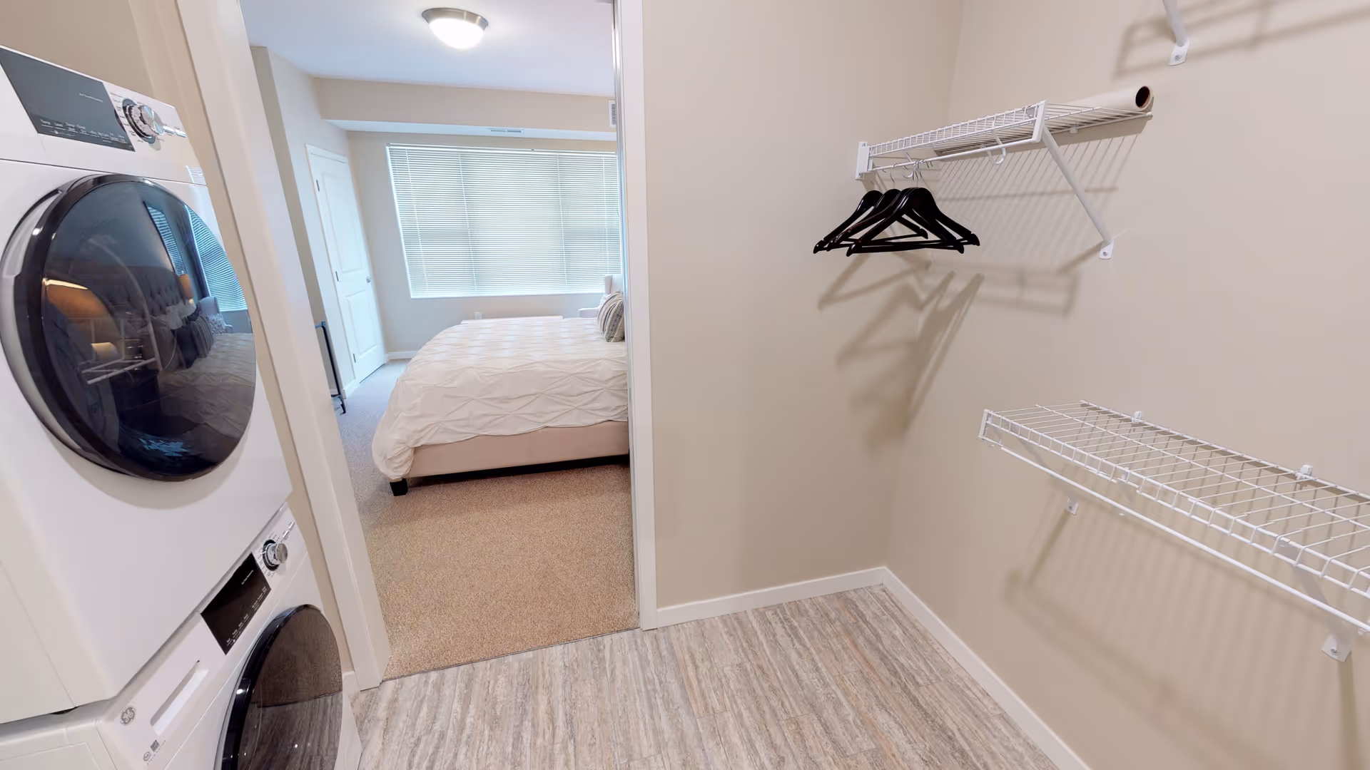 Laundry area with a stacked washer and dryer unit on the left, adjacent to a bedroom with a bed visible through an open doorway. The laundry room has light-colored walls, wood-patterned flooring, and wire shelving with black hangers on the right wall.