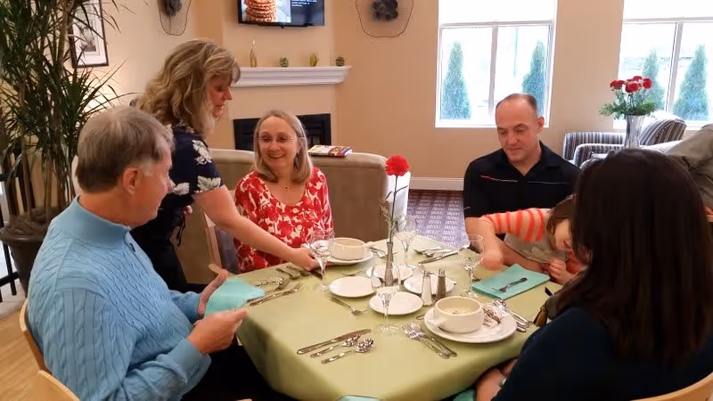 A group of five people sitting around a dining table in a cozy room. The table is set with plates, bowls, silverware, and glasses. A woman is serving a drink to an elderly woman in a red floral top. Other people at the table include an elderly man in a blue sweater, a man in a black shirt, a child in a striped shirt, and a woman with dark hair. The room has a fireplace, a TV mounted above it, and large windows letting in natural light.