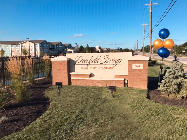 Entrance sign reading 'Deerfield Springs Resort Lifestyle Communities' on a brick monument with apartment buildings and balloons visible behind it.