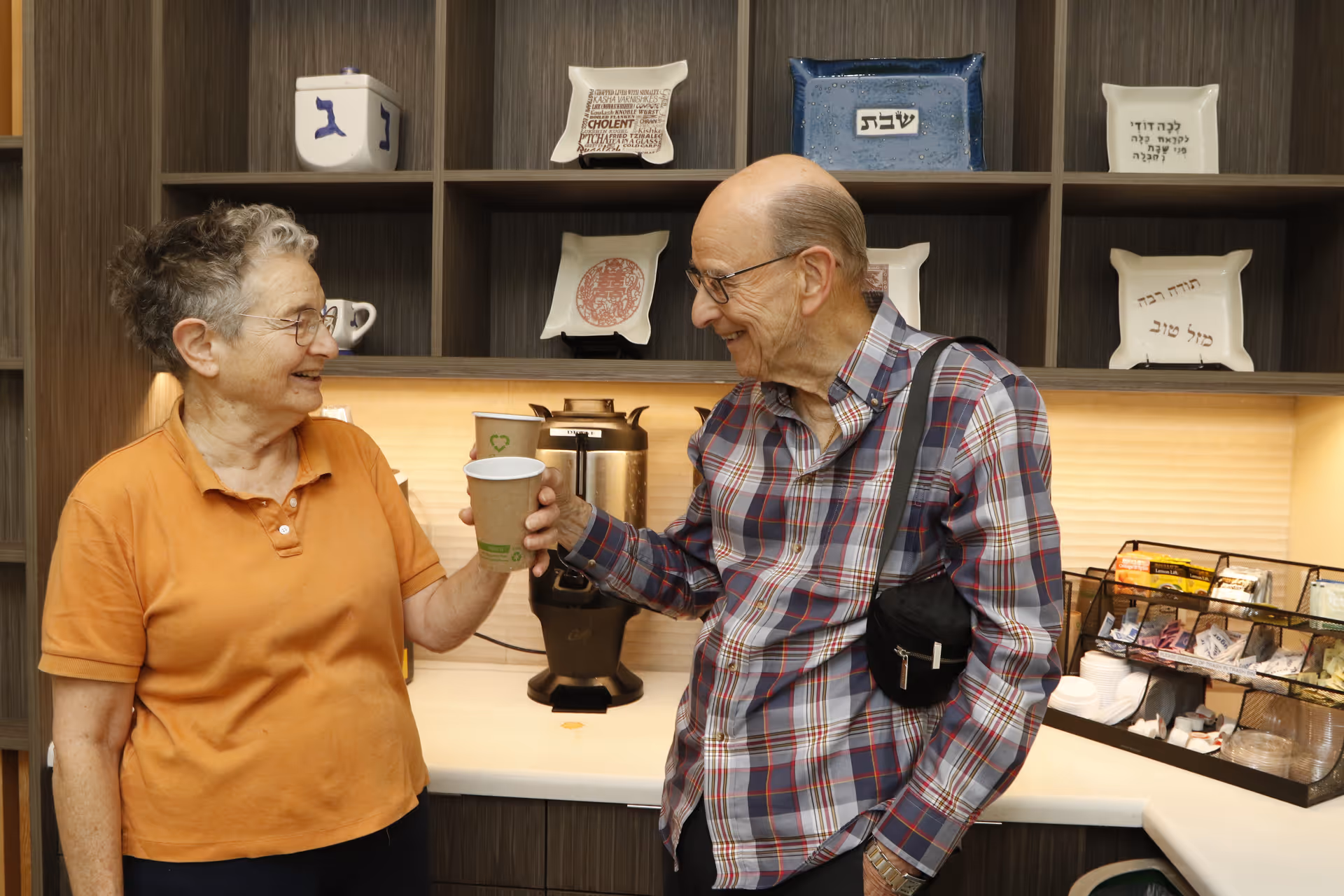 Two elderly individuals smiling and holding disposable cups in a kitchen or refreshment area with a coffee urn and a variety of condiments and supplies on the counter behind them.