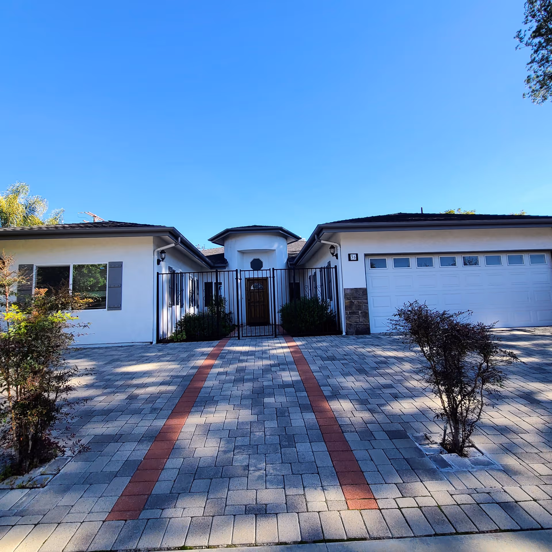Front exterior of a single-story white house with a gated entry, two-car garage, and a brick-patterned paver driveway under a clear blue sky.