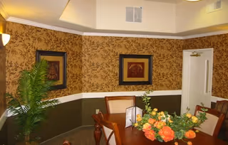 Dining area with a wooden table and chairs, a floral centerpiece, framed artwork, patterned wallpaper, and a potted plant.