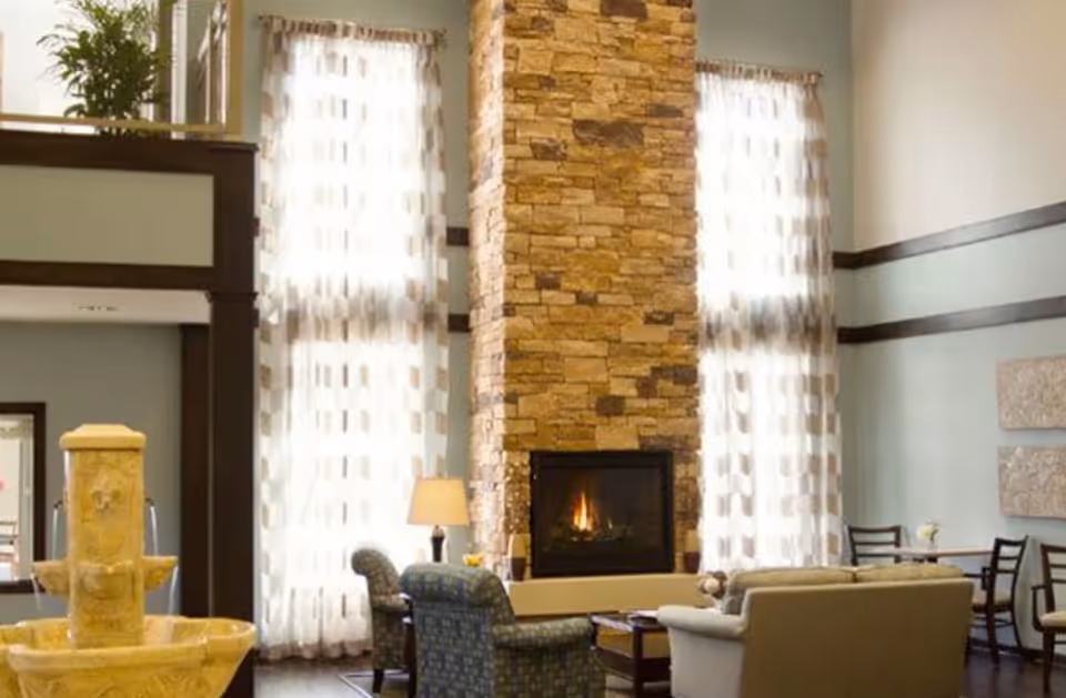 A cozy living room area with a tall stone fireplace in the center, flanked by two large windows with sheer patterned curtains. The room features comfortable armchairs and a sofa arranged around a coffee table, with a lamp on a side table. There is also a stone water fountain visible on the left side of the image.