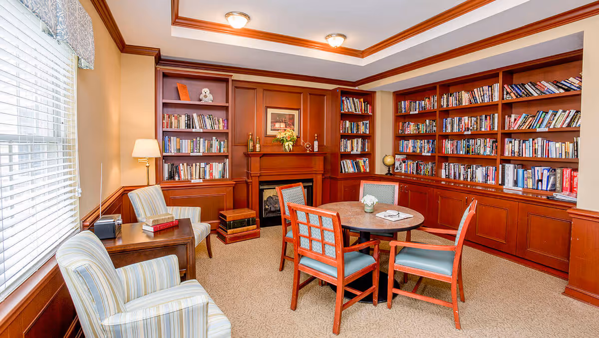 Cozy wood-paneled library room with built-in bookshelves, a round table with chairs, armchairs, and a fireplace.