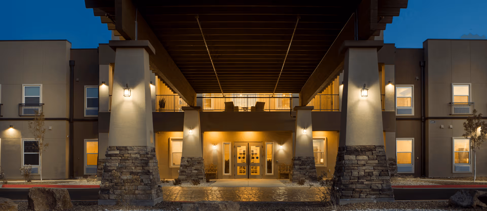 Front entrance of a two-story building at dusk, featuring illuminated pillars with stone bases, a covered walkway and an upper balcony.