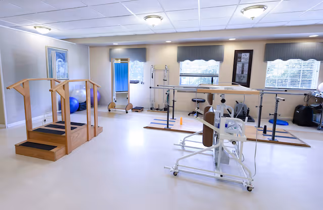 A bright rehabilitation therapy room with wooden parallel bars, exercise equipment, a mirror, and large windows with blinds letting in natural light.