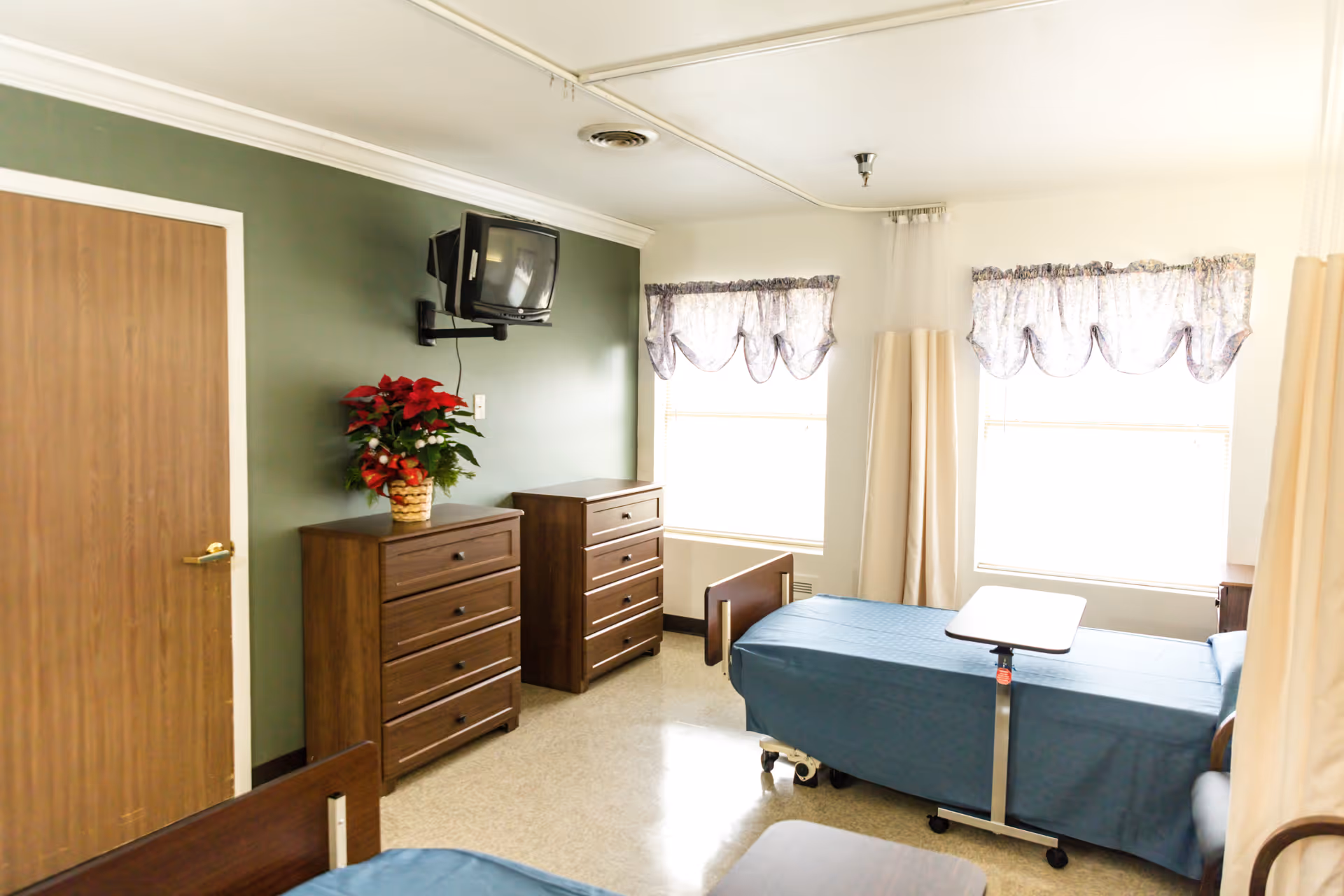 A bright room with two hospital beds covered in blue sheets, a small overbed table, two wooden dressers with a potted plant on one, a wall-mounted TV, two windows with floral valances, and beige curtains.