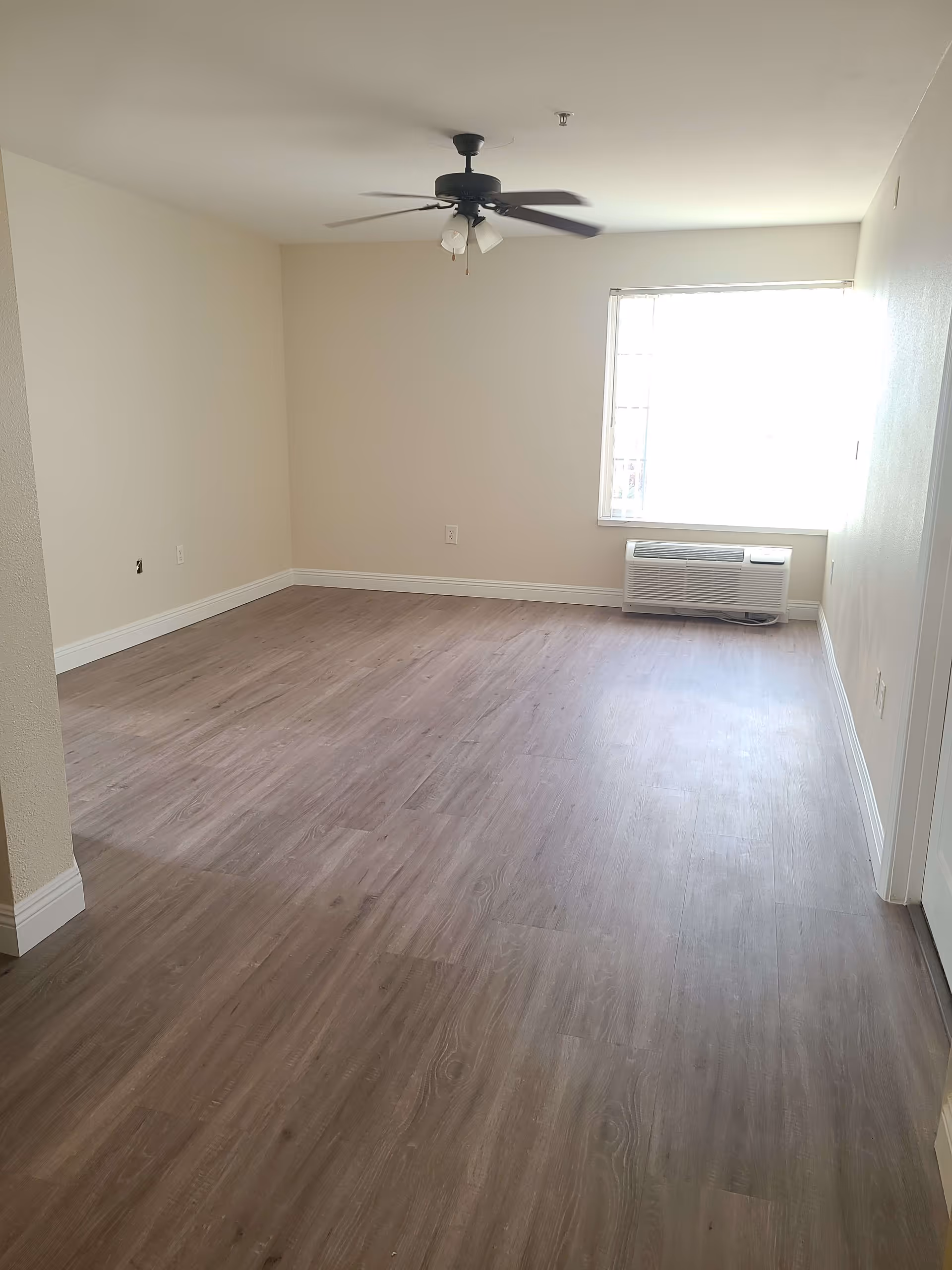 Empty room with light wood flooring, beige walls, a ceiling fan with lights, a window with blinds, and a wall-mounted air conditioning unit below the window.