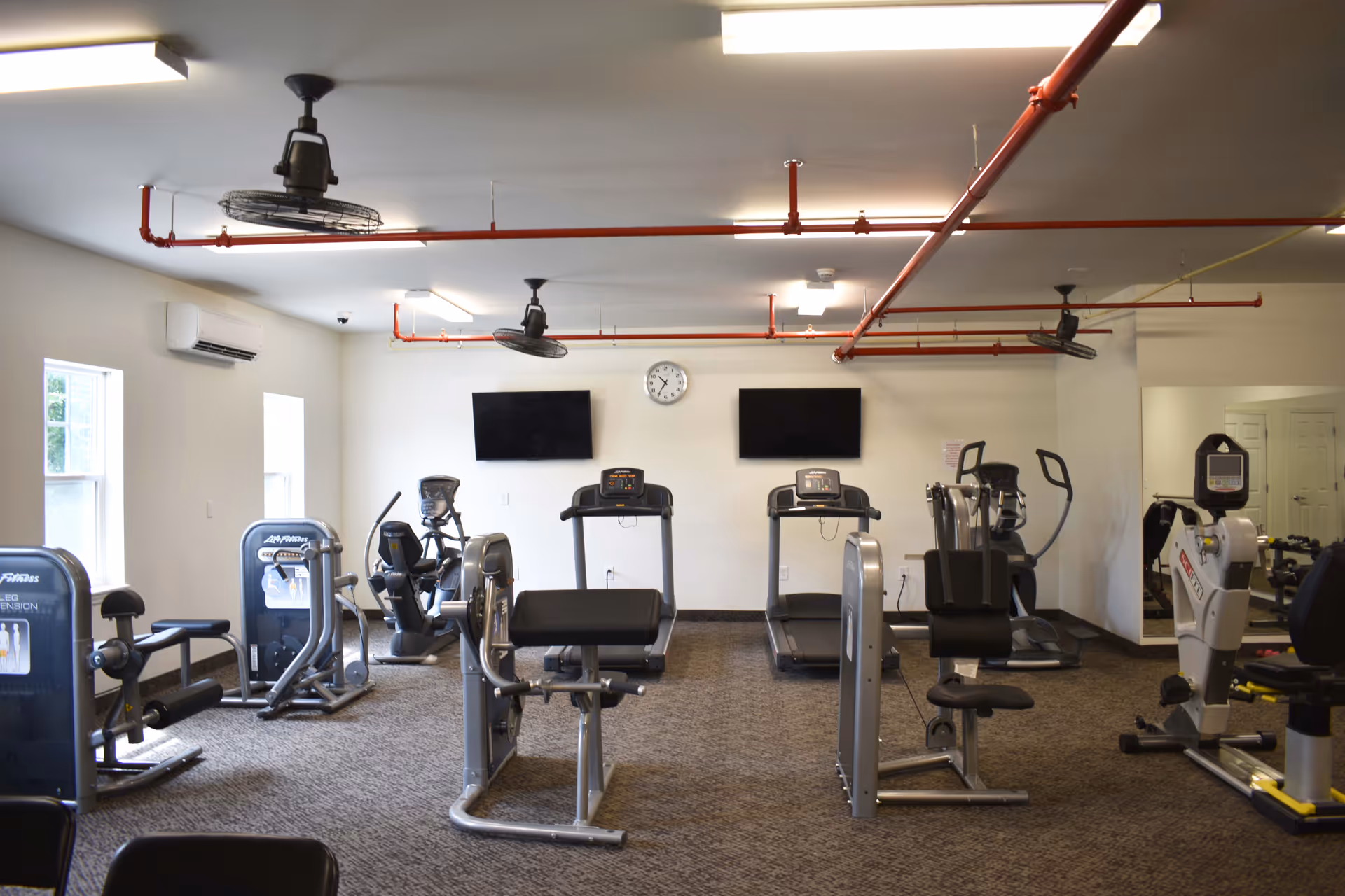 A fitness room with various exercise machines including treadmills, stationary bikes, and weight machines. The room has carpeted flooring, white walls, two windows on the left side, two wall-mounted TVs, a clock, and ceiling fans with exposed red piping.