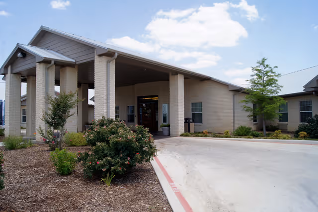 Covered entrance and driveway of a single-story beige brick nursing and rehabilitation building with landscaped shrubs under a blue sky.