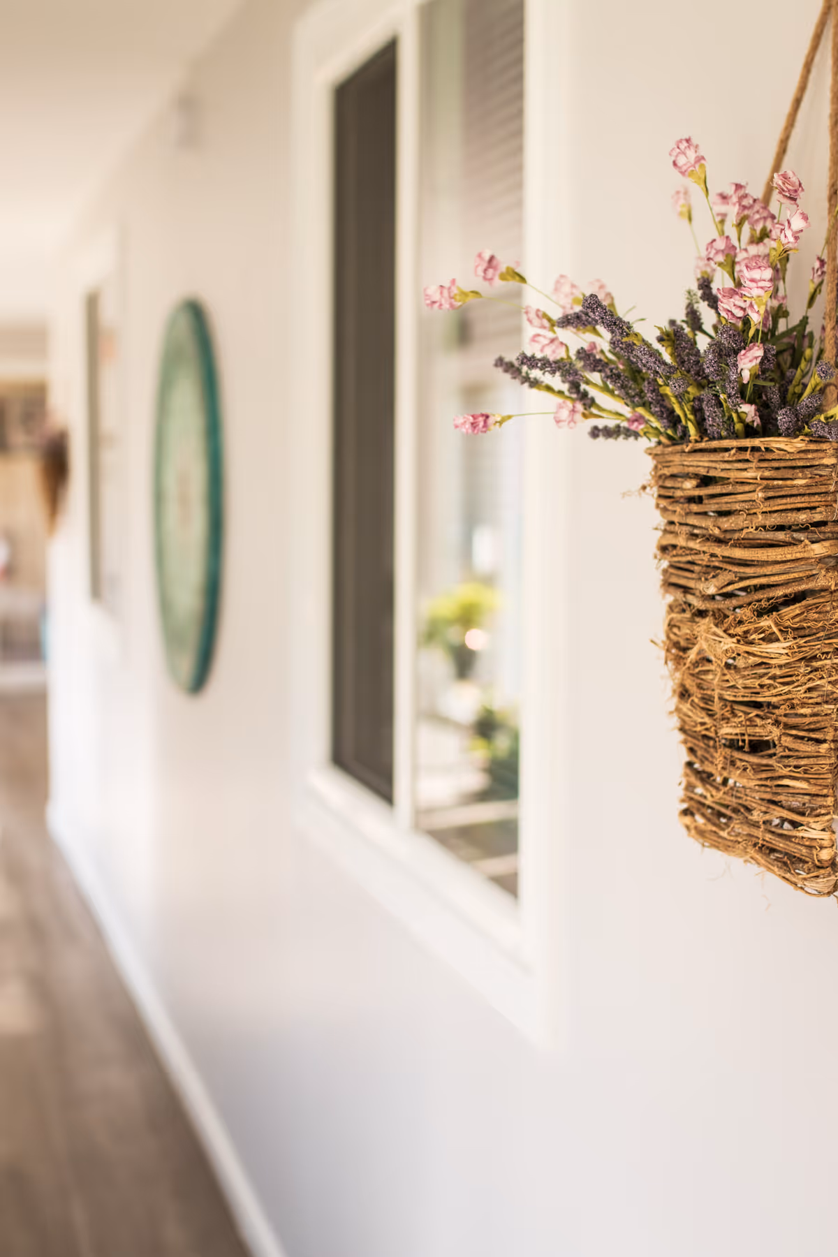 A bright interior hallway with a wicker hanging basket of flowers in the foreground and windows and wall decor blurred in the background.