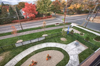 View of an outdoor fenced garden area with a curved concrete pathway, small landscaped sections with plants, and a basketball hoop. Beyond the fence is a sidewalk, a road, and trees with autumn foliage in red, yellow, and green colors.
