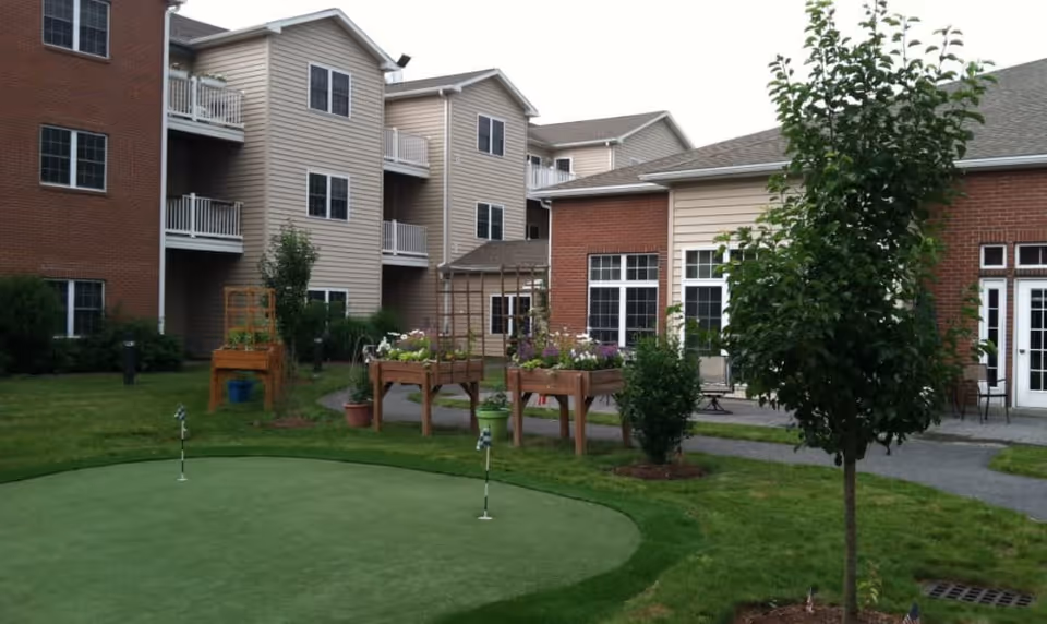 Outdoor garden area at Keystone Commons featuring a small putting green with two flags, raised garden beds with plants, a tree, and a multi-story residential building with balconies in the background.