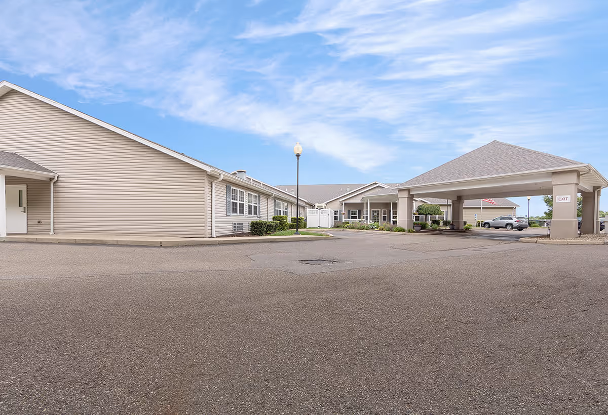 Exterior view of a single-story building with beige siding and a covered entrance area under a clear blue sky. There is a paved driveway and a few parked cars visible in the background.