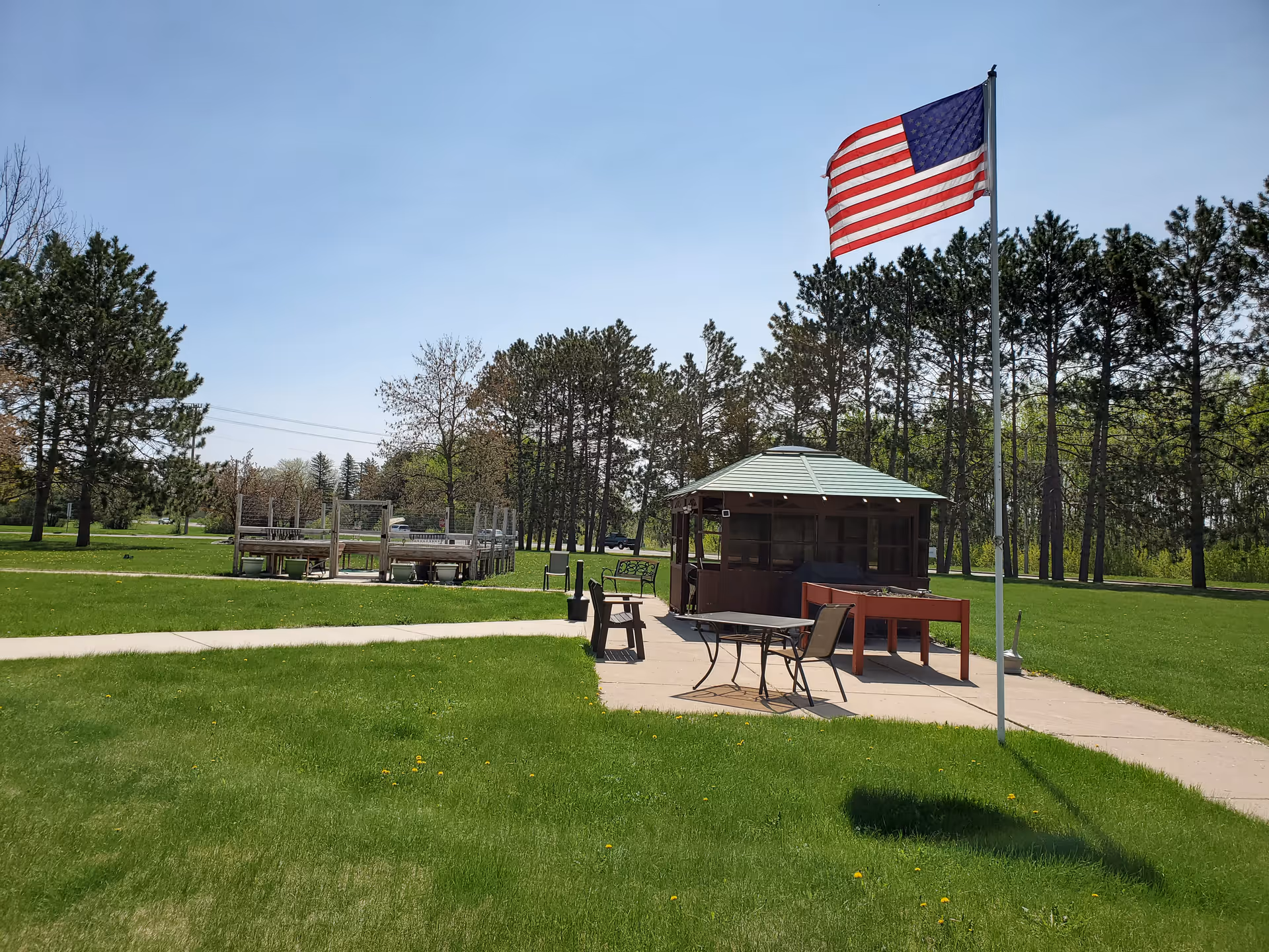 Outdoor community area with a gazebo, tables and chairs on a grassy lawn and a large American flag on a flagpole, surrounded by trees.