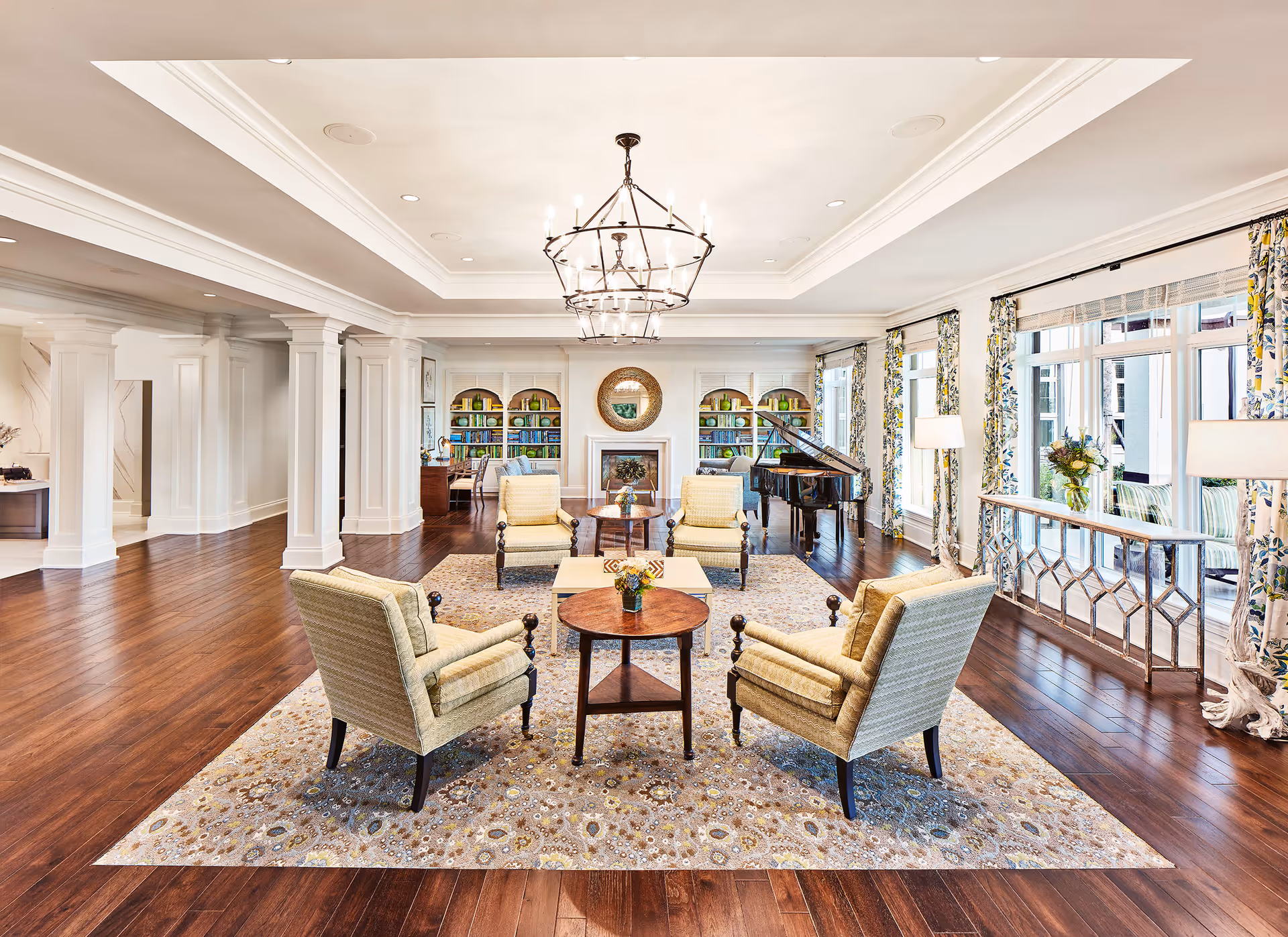 A spacious and elegant living room with four upholstered armchairs arranged around two round wooden tables on a patterned area rug. The room features dark hardwood floors, large windows with floral curtains, a grand piano, built-in bookshelves with decorative items, a fireplace with a round mirror above it, and two large chandeliers hanging from a tray ceiling.