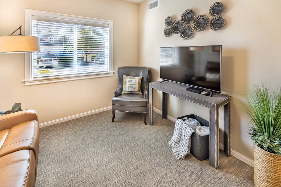 A cozy living room area with a large window with blinds, a leather armchair with a decorative pillow that says 'Thankful & Blessed', a modern console table with a flat-screen TV on top, and a basket with rolled blankets underneath. There is a floor lamp on the left and a tall plant in a woven basket on the right. Decorative wall art is mounted above the TV.