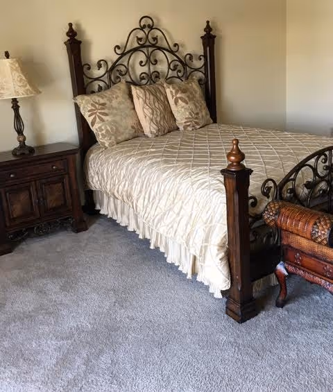 A bedroom with a decorative wrought iron and wooden bed frame, a cream-colored quilted bedspread, and three patterned pillows. Next to the bed is a wooden nightstand with a lamp featuring a beige lampshade. The floor is carpeted in a light gray color.