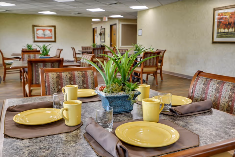 Dining area with a table set with yellow plates, matching mugs, glasses and brown napkins around a planter centerpiece, with more tables and chairs in the background.