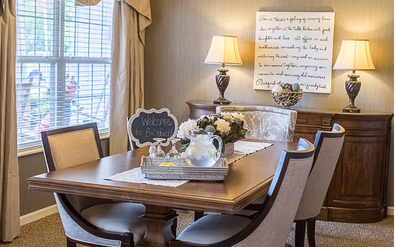 A warmly lit dining room with a wooden table set with a tray holding a pitcher of water, glasses, and a small chalkboard sign that reads 'Welcome to Bickford'. The table is surrounded by upholstered chairs. In the background, there are two table lamps on a wooden sideboard and a framed decorative sign on the wall. A window with blinds and curtains is on the left side.
