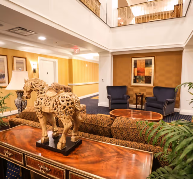 Ornate carved horse sculpture on a wooden console table in a furnished lobby seating area with armchairs and artwork.