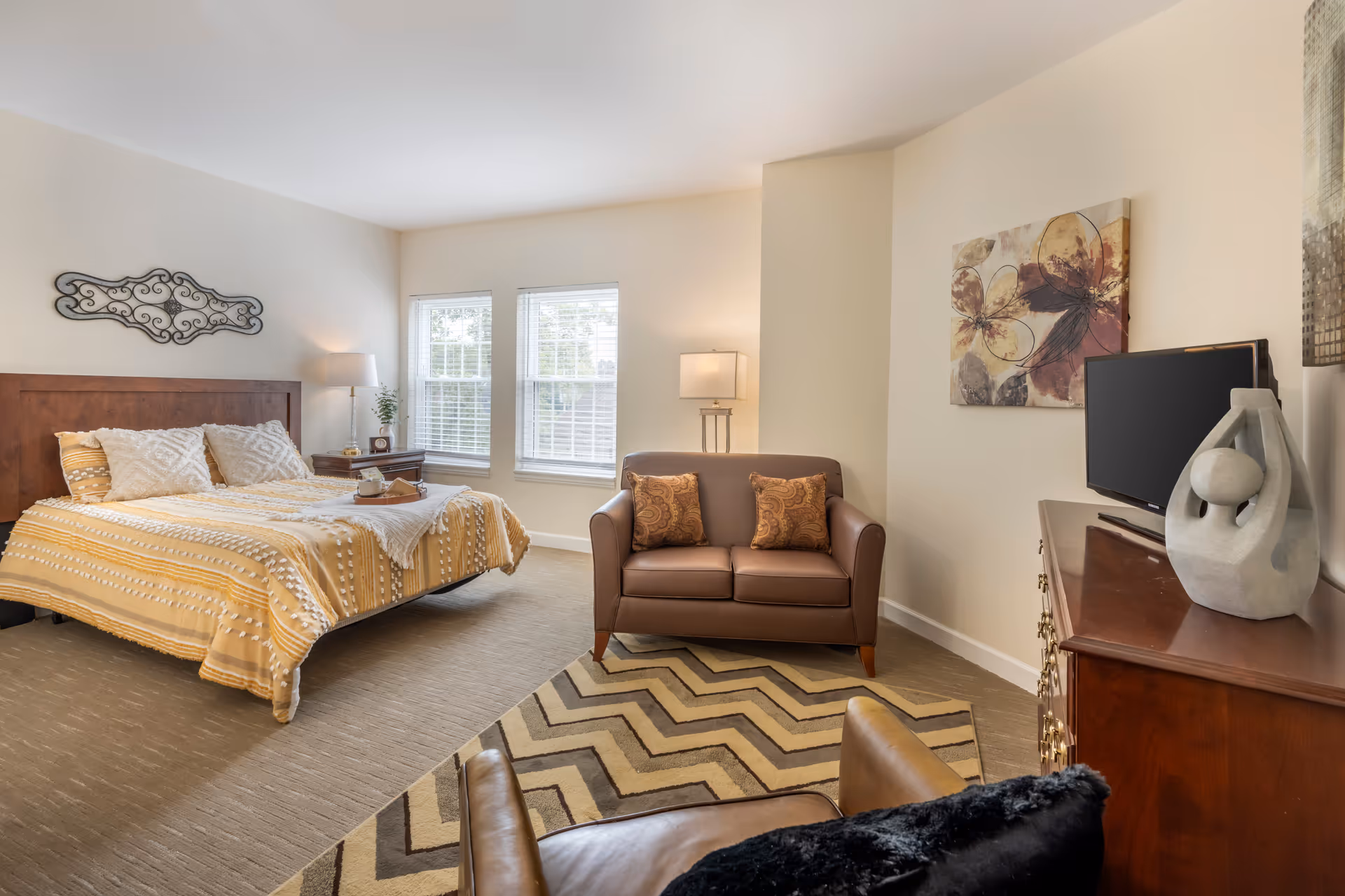 A cozy bedroom with a large bed covered in a yellow and white patterned blanket, two pillows, and a decorative wall piece above the headboard. Next to the bed is a wooden nightstand with a lamp and a small plant. The room also features a brown loveseat with two patterned cushions, a floor lamp behind it, a patterned rug, a wooden dresser with a TV on top, and a modern sculpture. Two windows with white blinds allow natural light into the room.