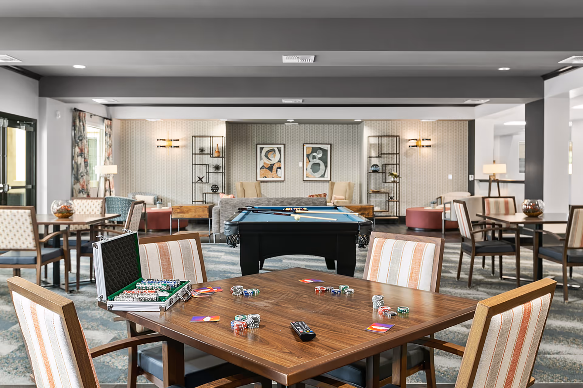 A spacious recreational room with a wooden table in the foreground set up for poker with poker chips, cards, and a remote control. Behind the table is a pool table with cues and balls arranged for a game. The room features comfortable seating areas with armchairs, sofas, and decorative shelving units against patterned wallpaper. The space is well-lit with ceiling lights and lamps, creating a welcoming atmosphere.