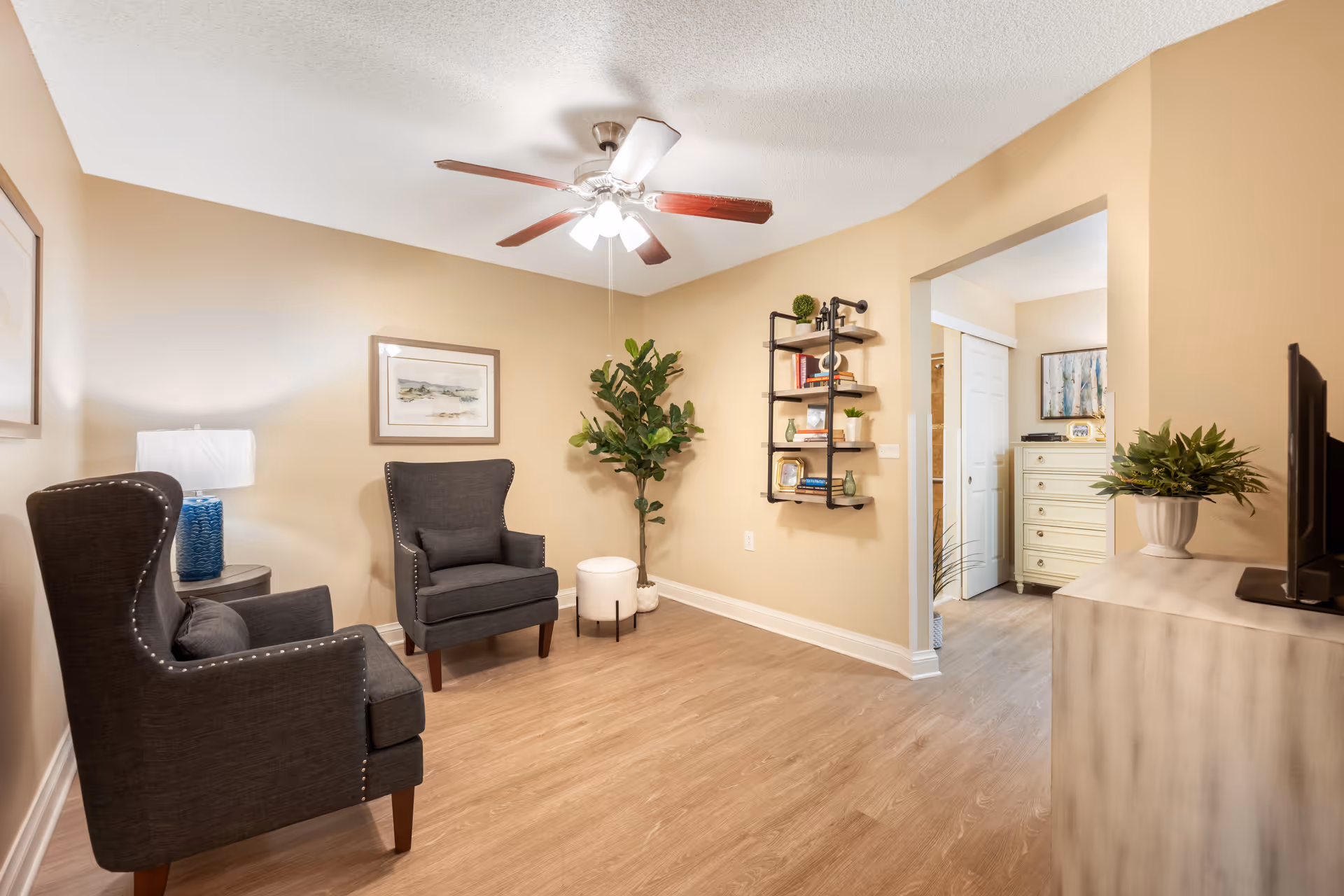 A cozy living room area with two dark gray armchairs, a small round side table with a blue lamp, a ceiling fan with lights, a potted plant, and a wall-mounted shelf holding books and decorative items. The room has light beige walls and wood flooring, with an open doorway leading to another room with a dresser and artwork.