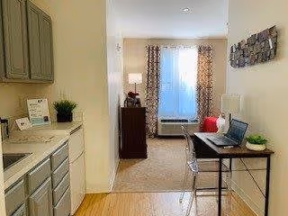 View of a small kitchenette area with cabinets, a sink, and countertop on the left, leading into a living space with a window covered by patterned curtains. A desk with a laptop and a clear chair is positioned on the right side of the image, and a small plant and lamp are visible in the living area.