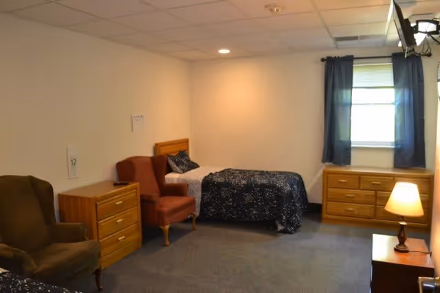 A simple furnished bedroom with a single bed, two armchairs, dressers, a lamp, and a window with blue curtains.