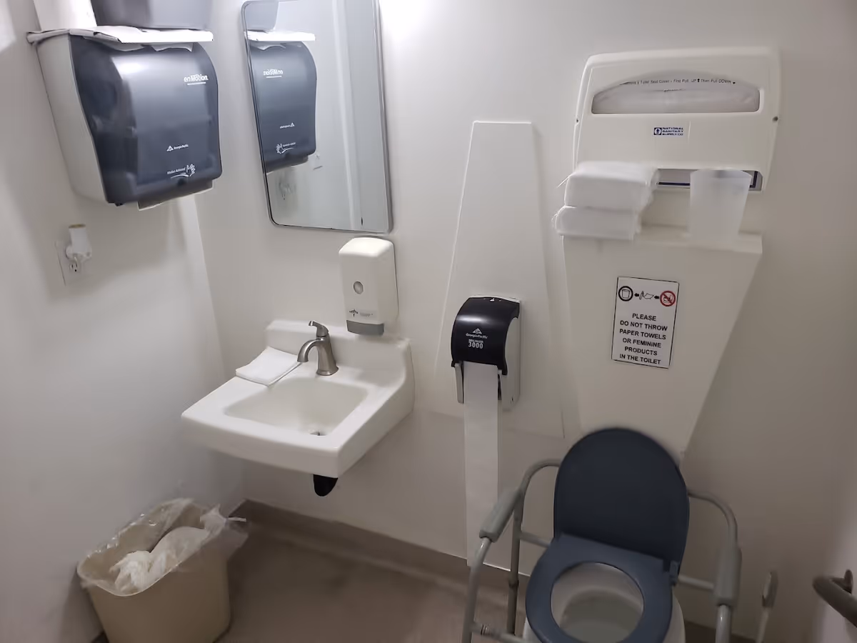 Small institutional bathroom with a wall-mounted sink and mirror, multiple paper towel dispensers, a wall-mounted toilet paper dispenser, and a raised commode chair.