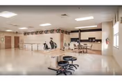 A spacious room with a clean, polished floor featuring a kitchen area with cabinets, a microwave, and a refrigerator. There are tables and chairs arranged near the kitchen, and stools around a counter in the foreground. The walls are decorated with a mural and some framed pictures. The ceiling has recessed lighting.