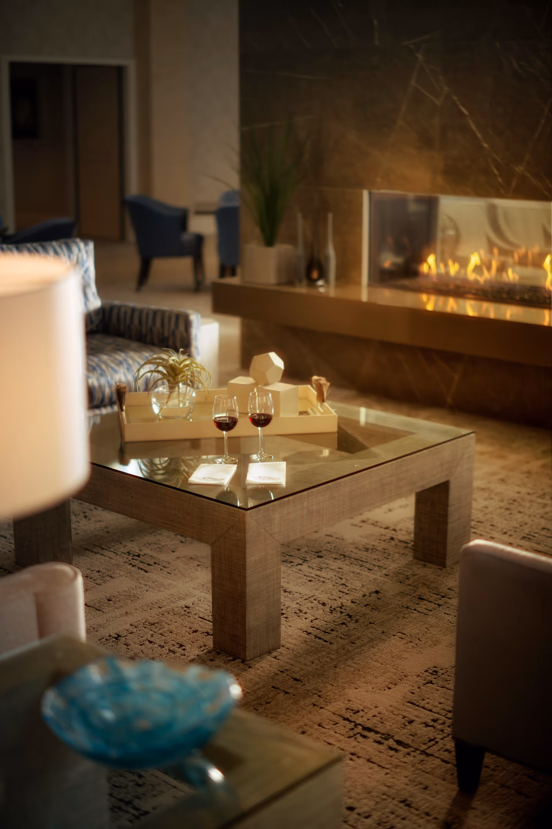A cozy living room area with a glass-top wooden coffee table holding two glasses of red wine and a decorative tray with geometric objects and a small plant. In the background, there is a modern fireplace with a warm fire burning and some blue upholstered chairs near a doorway.