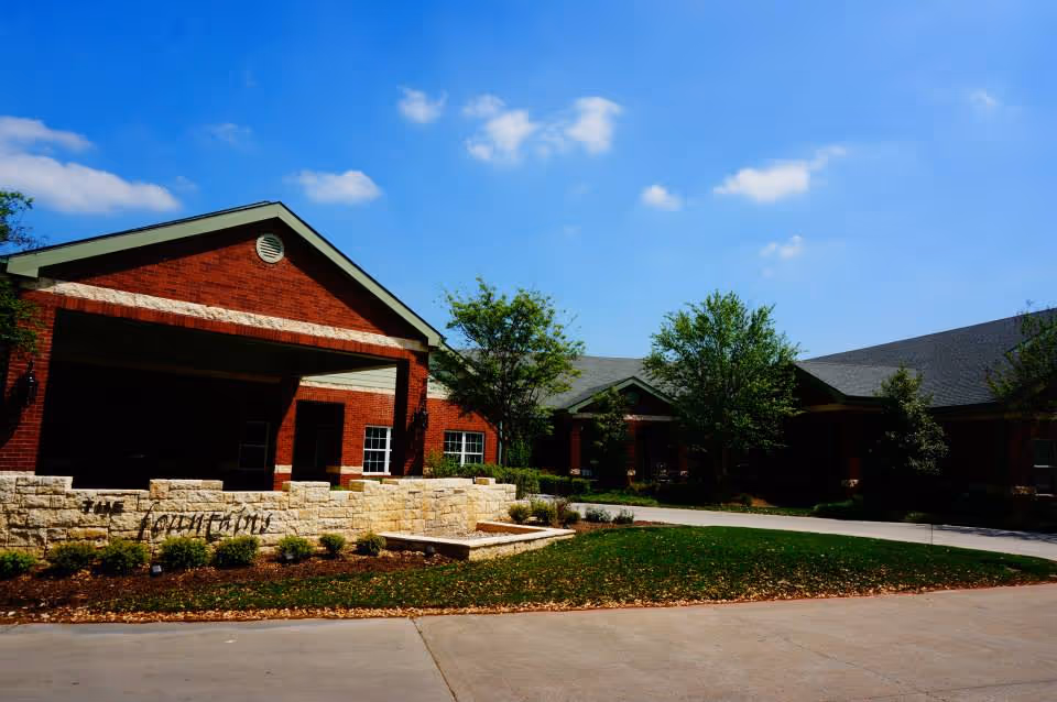 Exterior view of a single-story brick building with a covered entrance, surrounded by landscaped greenery and trees under a blue sky with scattered clouds.