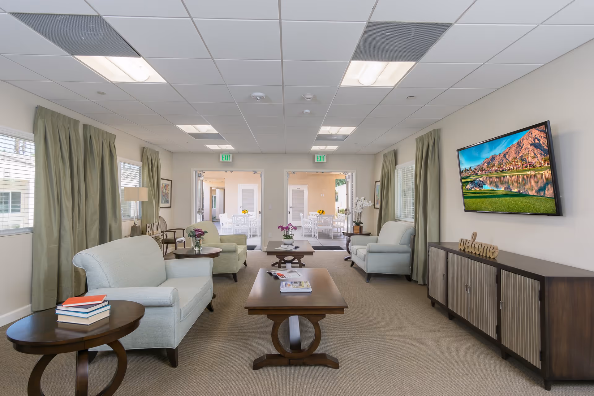 Well-lit communal living room with light blue armchairs, wooden coffee tables, a wall-mounted TV, and a doorway leading to an outdoor seating area.