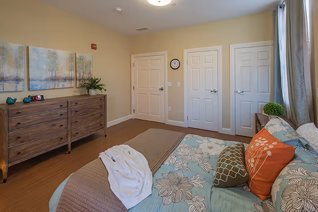 A bedroom with a bed covered in floral and patterned bedding, several pillows, and a folded white robe on top. There is a wooden dresser with decorative items and a plant on it against one wall. Three white doors and a wall clock are visible on the opposite wall. The room has beige walls and wood flooring.