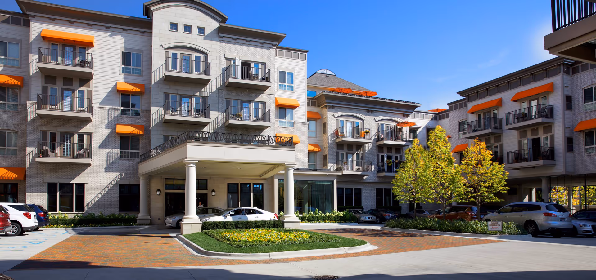 Front exterior of a multi-story senior living building with balconies, orange awnings, a covered entrance, parked cars, and landscaping.