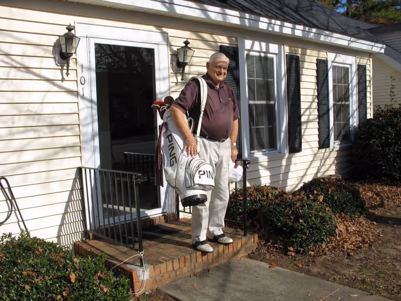 An elderly man standing outside a light-colored house with black shutters, holding a white golf bag with clubs. The house has a small brick porch with a black metal railing and two wall-mounted lanterns beside the door.