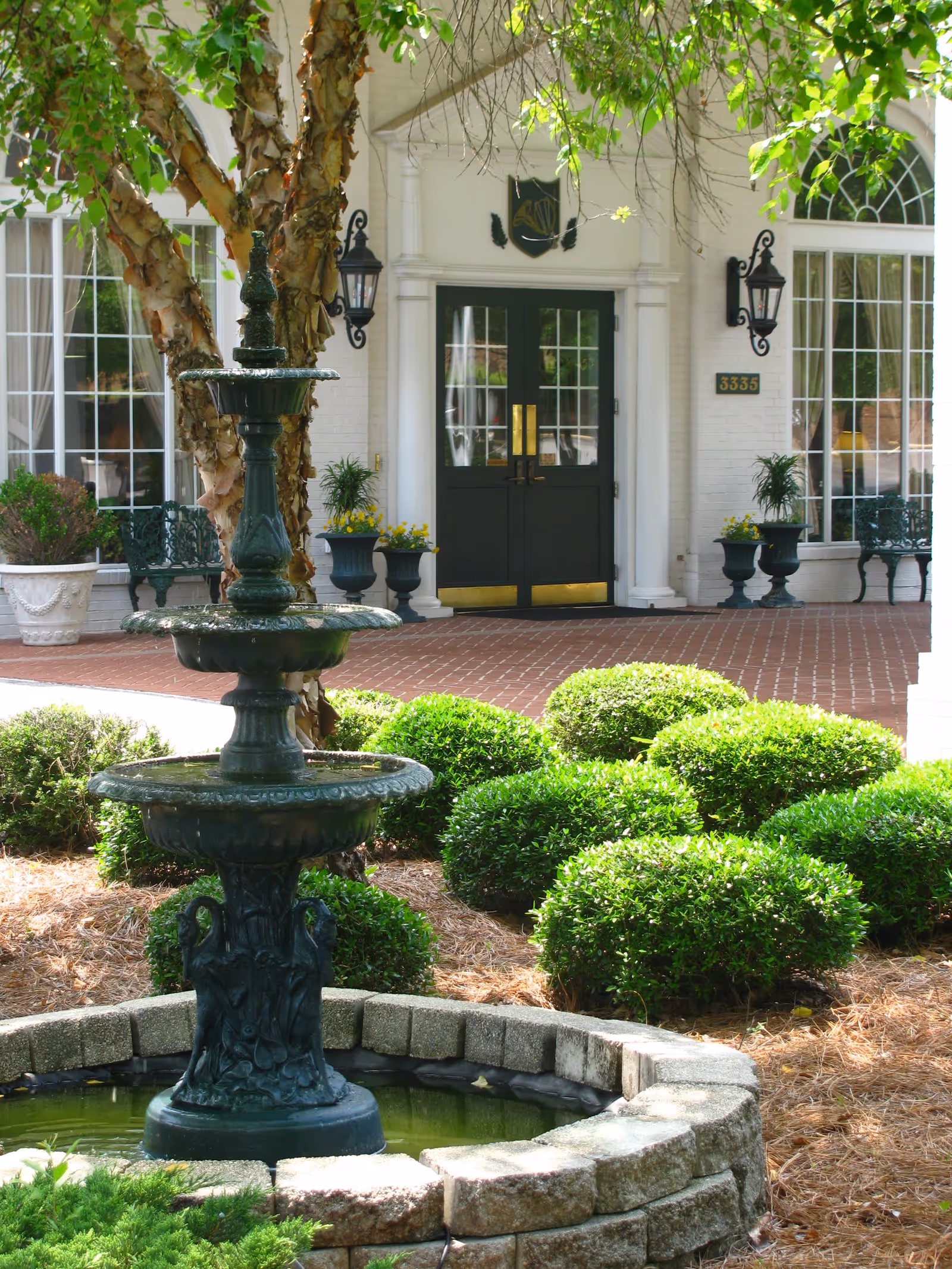 Outdoor view of the entrance to Cumberland Village featuring a decorative three-tiered fountain in the foreground, neatly trimmed bushes, a tree, and a brick walkway leading to double black doors with gold handles. The building facade is white with large windows and black wall-mounted lanterns on either side of the door.