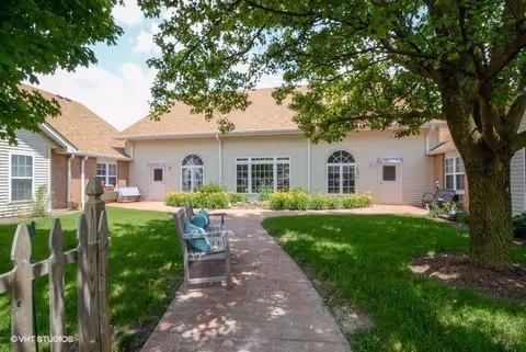 Outdoor courtyard area of a senior living facility with a paved walkway, green grass, benches with cushions, trees providing shade, and a building with large windows and doors in the background.