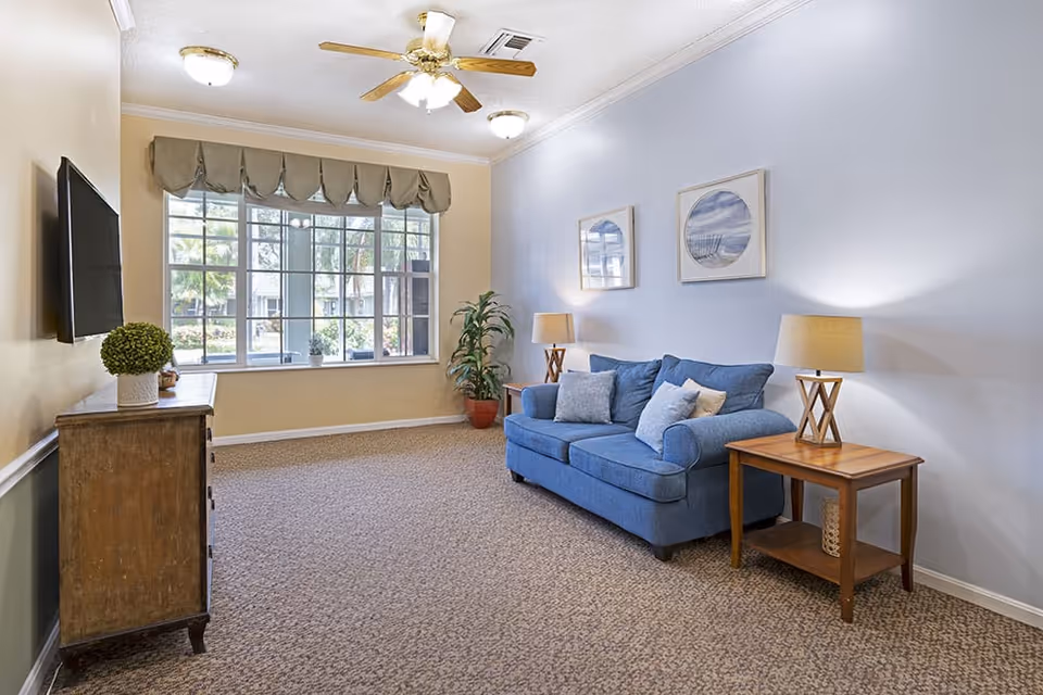 Bright sitting area with a blue sofa, side tables and lamps, a wall-mounted TV and a large window.