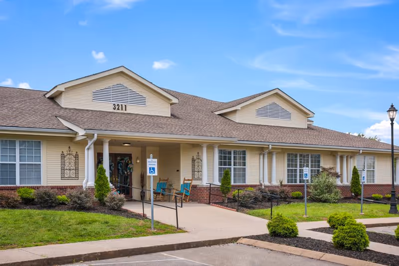 Exterior view of a single-story senior living facility building with beige siding and a brown shingled roof. The building has large windows, a covered entrance with two blue rocking chairs, and a ramp with handrails leading to the door. The address number 3211 is displayed above the entrance. There are small bushes and landscaped greenery around the building, a parking area with handicap parking signs, and a lamppost on the right side. The sky is clear and blue with a few clouds.