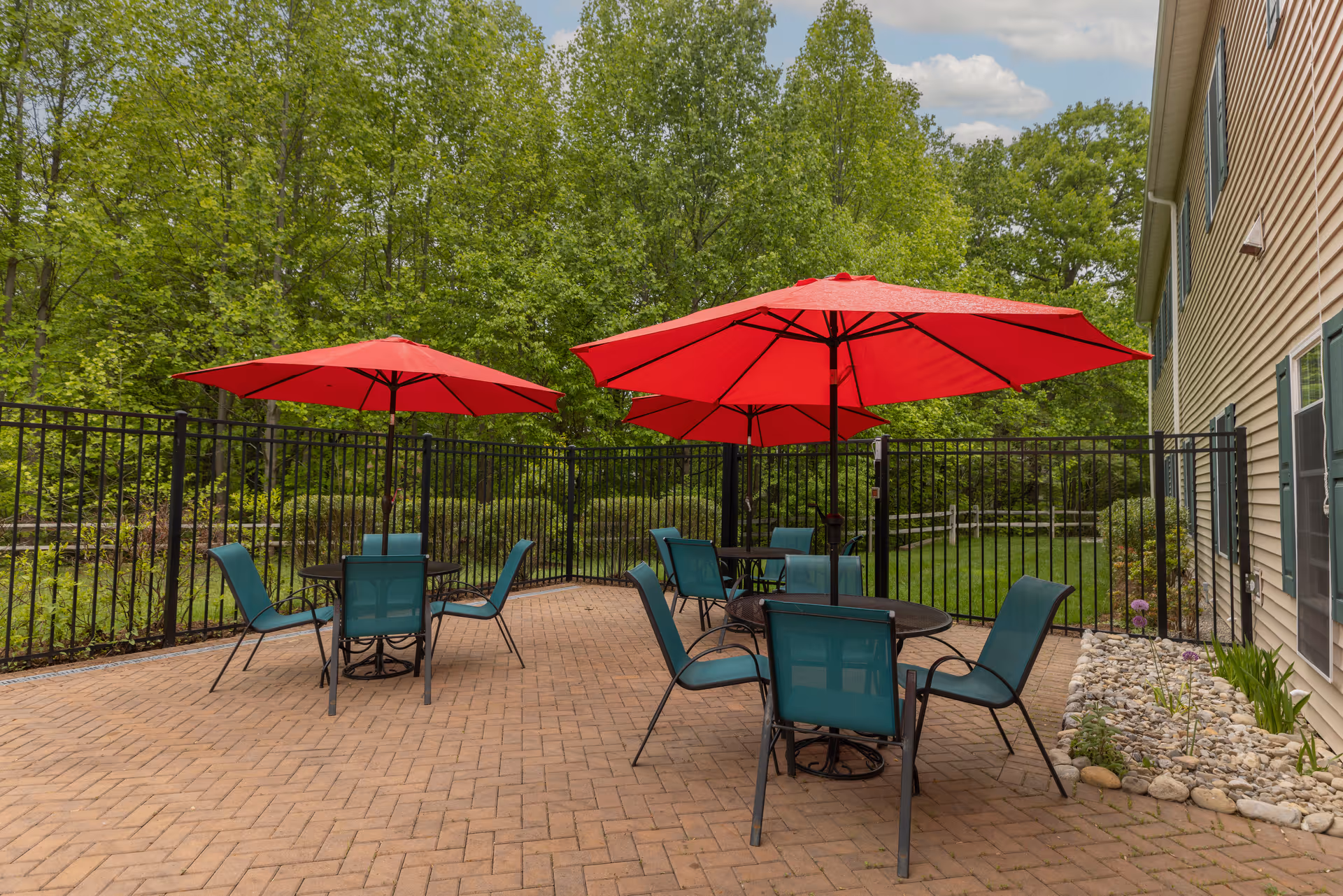 Outdoor patio with red umbrellas and teal chairs arranged around tables on a brick-paved area next to a fenced yard and trees.